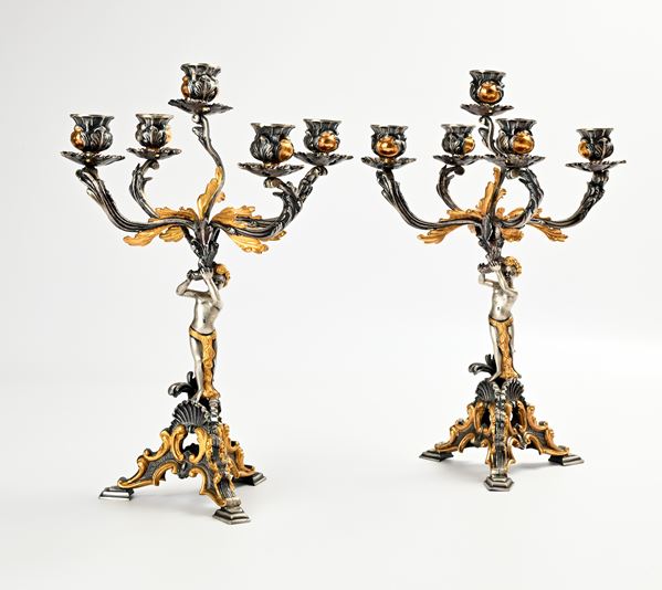 Coppia di candelabri in lega metallica a patina argentata e in parte dorata, cinque luci con figure di amorino su base tripode